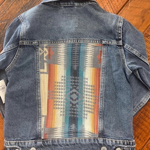 May include: A blue denim jacket with a colorful patterned fabric panel on the back. The pattern features geometric shapes in shades of blue, orange, yellow, and red.