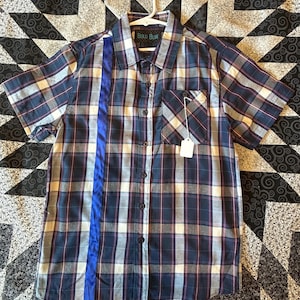 May include: A blue, white and red plaid short-sleeved button-down shirt with a blue stripe down the front. The shirt has a pocket on the left chest and a tag that says "Bold Blue".