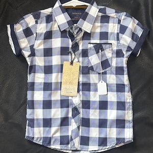 May include: A short-sleeved button-down shirt in a blue and white checkered pattern. The shirt has a small pocket on the chest and a collar. The tag reads "NOMAD". The shirt is displayed on a white hanger.
