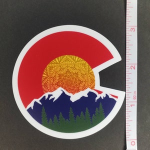 May include: A round sticker with a white border, featuring a stylized depiction of the Colorado state flag. The flag is represented by a red circle with a yellow mandala-like sun design and blue mountains with green trees at the base.