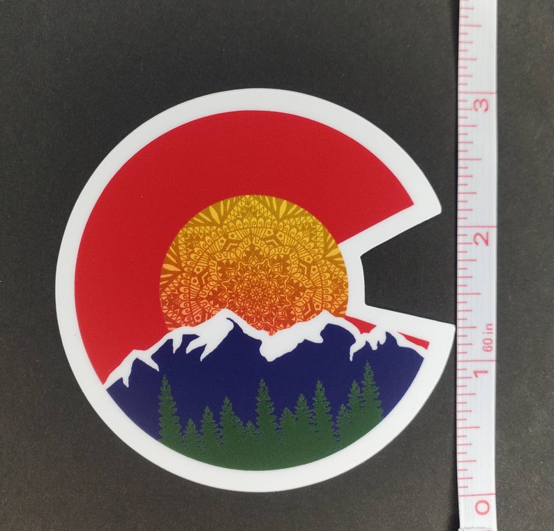 May include: A round sticker with a white border, featuring a stylized depiction of the Colorado state flag. The flag is represented by a red circle with a yellow mandala-like sun design and blue mountains with green trees at the base.