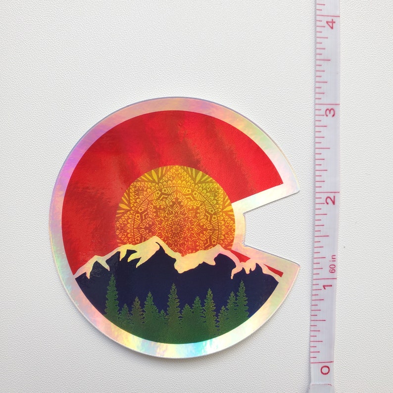 May include: A holographic sticker featuring a red, yellow, and orange sunset over a mountain range with a mandala design. The sticker is shaped like the state of Colorado.
