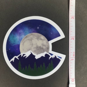May include: A sticker featuring a stylized outline of the state of Colorado with a full moon, mountains, and a starry night sky.