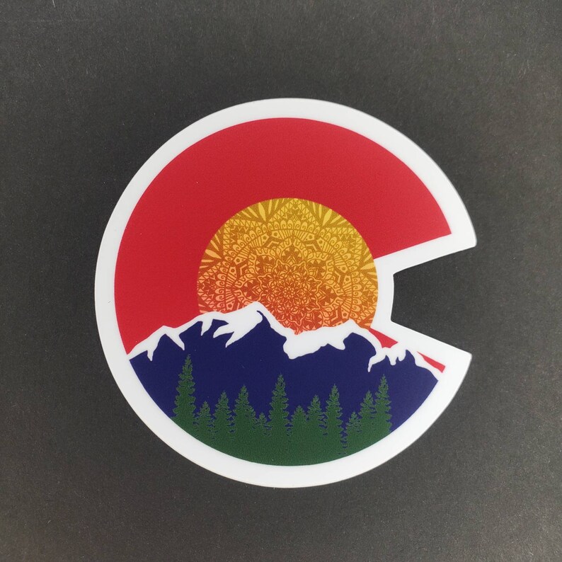 May include: A white sticker with a red, yellow, and blue design. The design features a stylized mountain range with a mandala-style sun rising behind it. The sticker is shaped like the state of Colorado.
