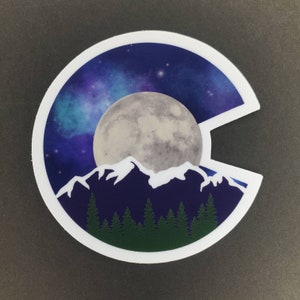 May include: A white sticker with a blue, purple, and white starry sky background. A large, gray moon is in the center of the sticker. A white mountain range is in front of the moon, and a dark blue mountain range is behind the moon. A row of dark green trees is at the bottom of the sticker.
