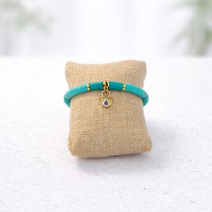 May include: A turquoise beaded bracelet with gold detailing and a gold Hamsa hand charm with a blue centre. The bracelet is displayed on a small, square, tan cushion.