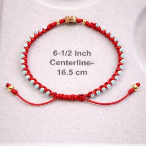 May include: Red macrame bracelet with light blue beads and gold accents. The bracelet is 16.5 cm in length. The bracelet has a sliding knot closure for an adjustable fit. The bracelet is displayed on a light grey surface.
