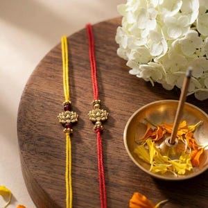 May include: Two decorative bracelets featuring thin yellow and red threads, each adorned with a central gold-toned lotus flower charm and small dark red beads, presented as a pair of traditional wrist ornaments for festive occasions.