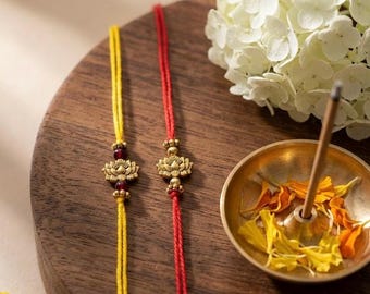 May include: Two decorative bracelets featuring thin yellow and red threads. Each bracelet has a central gold-toned metal lotus flower charm. The yellow thread bracelet includes small dark red beads flanking the lotus charm, while the red thread bracelet features the gold lotus charm centred between small gold-toned metal beads.