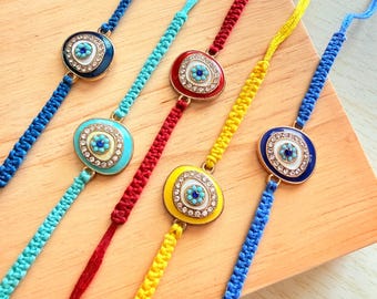 May include: Five adjustable bracelets featuring woven cords in blue, turquoise, red, yellow, and royal blue. Each bracelet centres on an oval charm with a central blue eye motif, surrounded by white enamel and a border of small clear crystals.