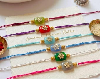 May include: Six handmade macrame rakhis featuring rectangular beads in green, red, light blue, medium blue, dark green, and grey. Each bead is centred with a gold-toned floral accent set with clear stones on thin, colourful braided cords.
