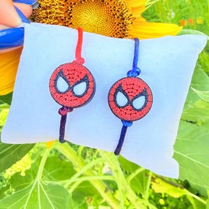 May include: Two circular bracelets featuring a red and black textured mask design with white eye accents. One bracelet has a red cord, and the other has a blue cord. Both are mounted on a white rectangular display cushion for clear presentation.