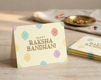 May include: A cream-coloured folded greeting card with the text HAPPY RAKSHA BANDHAN! in brown, decorated with small, colourful mandala-style floral patterns in shades of blue, orange, yellow, purple, and green, with a stack of identical cards nearby.