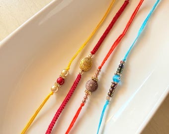 May include: Four bracelets made of thin yellow, red, orange, and light blue cord. The yellow bracelet features two cream beads and one red bead. The red bracelet has a gold flower charm. The orange bracelet has a patterned brown bead. The light blue bracelet features metallic beads and a small blue evil eye bead.