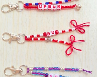May include: Three beaded keychains with silver clasps. One has red, white, and blue beads with "UPENN". Another has red and white beads with "BU" and a heart charm. The third has purple and blue beads with "NU".
