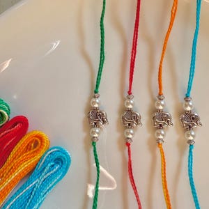 May include: Four bracelets featuring green, red, orange, and blue twisted threads. Each bracelet is centered with a silver-toned metal elephant charm flanked by two white faux pearls and small silver-toned decorative spacer beads.