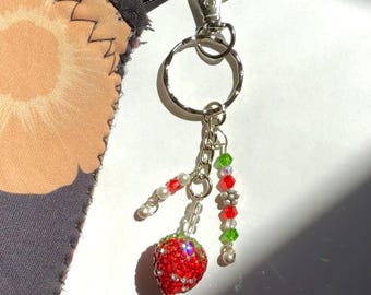May include: A silver-toned metal keyring featuring a strawberry charm covered in red crystals. The keyring includes two dangling beaded strands with red, green, and white beads, attached to a circular ring and a lobster claw clasp.