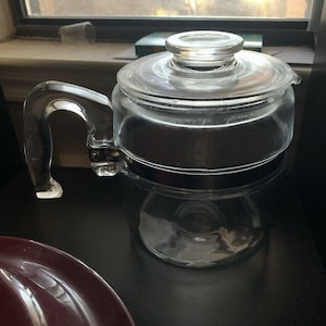 May include: A clear glass teapot with a curved handle and a matching lid. The teapot is sitting on a dark surface, reflecting its shape. The handle is thick and sturdy, and the lid has a round knob for easy removal. The teapot appears to be empty.