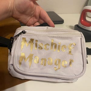 May include: A light purple fanny pack with the words "Mischief Manager" in gold lettering. The bag has a black strap and zipper. A small red and white crafting tool is in the background.