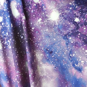 Pode incluir: Tecido roxo, azul e branco com estampa de gal&aacute;xia. O tecido tem uma superf&iacute;cie texturizada e est&aacute; dobrado no centro.