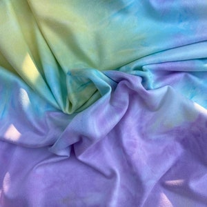 Pode incluir: Um close-up de um tecido tie-dye em tons de amarelo, azul e roxo. O tecido é macio e enrugado, criando um visual texturizado.