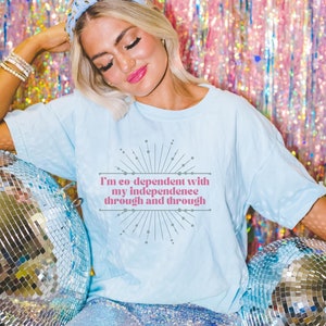 May include: A woman wearing a light blue t-shirt with the text "I'm so dependent with my independence through and through" in pink lettering. She is sitting on a white surface with two silver disco balls.