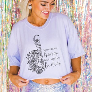 May include: A light purple cropped t-shirt with a black and white graphic of a skeleton with flowers and the text "You collected bones but I buried the bodies".