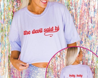 Puede incluir: Una camiseta recortada lavanda con el texto "the devil said hi" en rojo con una cola de diablo roja. La parte posterior de la camiseta tiene el texto "baby, baby look at me now" en rojo.