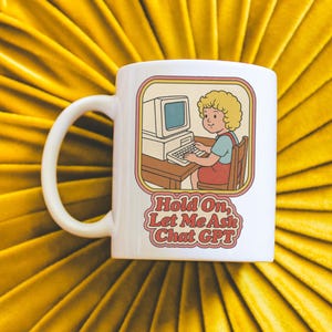 May include: White ceramic mug featuring a retro illustration of a person at a computer, with the text "Hold On, Let Me Ask Chat GPT." The design is framed in a vintage style, with a yellow, orange, and brown color scheme.