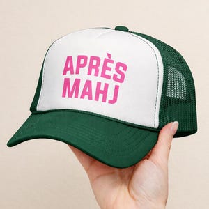 Op de afbeelding: Groene en witte truckerpet met de roze tekst "APR&Egrave;S MAHJ". De pet heeft een gebogen klep en een mesh achterkant. De pet wordt door een hand omhoog gehouden.