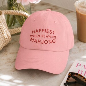 May include: A light pink baseball cap with the text "HAPPIEST WHEN PLAYING MAHJONG" embroidered in a darker pink. The cap is sitting on a table next to a book, sunglasses, and a drink.