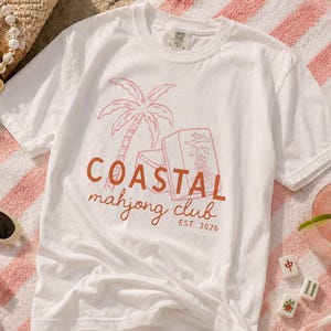 Puede incluir: Camiseta blanca con un gr&aacute;fico coral y rosa de una palmera, fichas de mahjong y el texto "COASTAL mahjong club EST. 2026." La camiseta est&aacute; sobre una toalla a rayas rosas y blancas, con gafas de sol y una bebida.