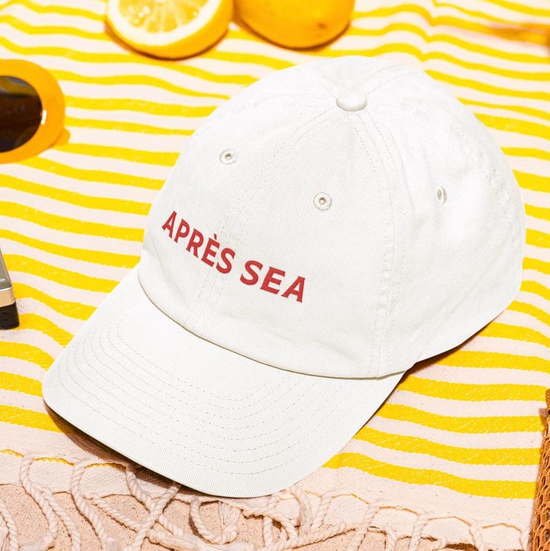 May include: A cream-coloured baseball cap with the words "APR&Egrave;S SEA" in red lettering. The cap is on a yellow and white striped towel, with orange sunglasses and a lemon in the background.