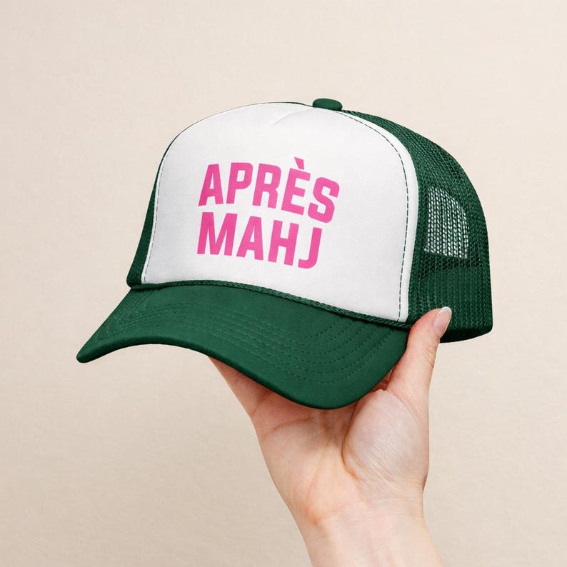 Op de afbeelding: Groene en witte truckerpet met de roze tekst "APR&Egrave;S MAHJ". De pet heeft een gebogen klep en een mesh achterkant. De pet wordt door een hand omhoog gehouden.