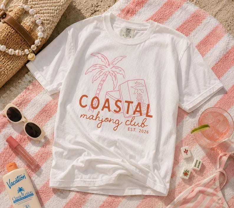 Puede incluir: Camiseta blanca con un gr&aacute;fico coral y rosa de una palmera, fichas de mahjong y el texto "COASTAL mahjong club EST. 2026." La camiseta est&aacute; sobre una toalla a rayas rosas y blancas, con gafas de sol y una bebida.