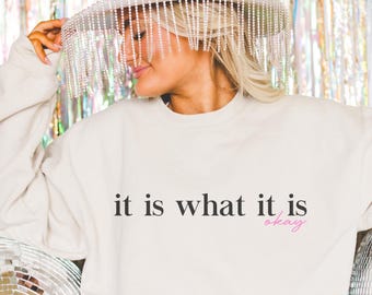 May include: A white sweatshirt with the text "it is what it is okay" printed on the front. The sweatshirt is being worn by a person wearing a white hat with a beaded fringe.