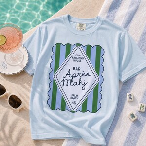 May include: Light blue t-shirt with a graphic that reads "The Mahjong House Bar Apr&egrave;s Mahj Palm Beach FL 2026". The shirt is next to a cocktail, sunglasses, and Mahjong tiles on a striped towel.