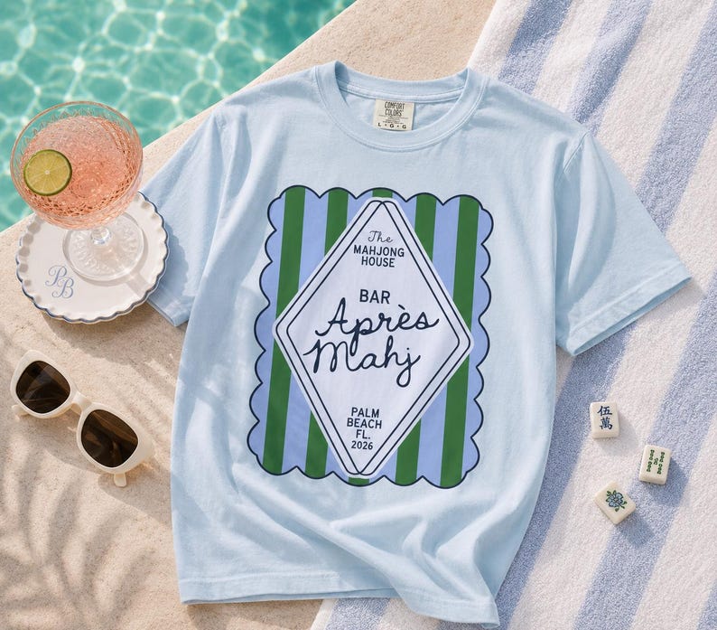 May include: Light blue t-shirt with a graphic that reads "The Mahjong House Bar Apr&egrave;s Mahj Palm Beach FL 2026". The shirt is next to a cocktail, sunglasses, and Mahjong tiles on a striped towel.