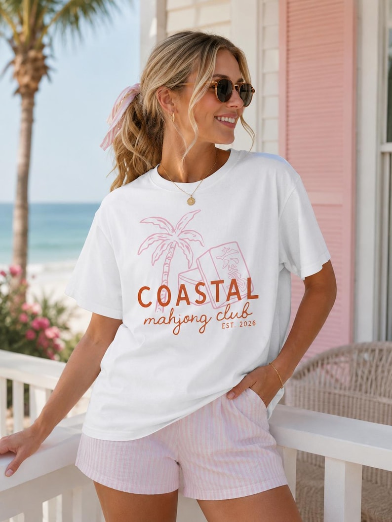 Puede incluir: Camiseta blanca con una palmera rosa y el texto "COASTAL mahjong club EST. 2026." La camiseta se combina con pantalones cortos a rayas rosas y blancas. La imagen se toma al aire libre en un d&iacute;a soleado.