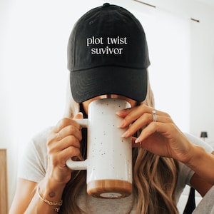 May include: Black baseball cap with the text "plot twist suvivor" in white. A white mug with black speckles and a wooden base is held up to the face. The person is wearing a bracelet and ring.