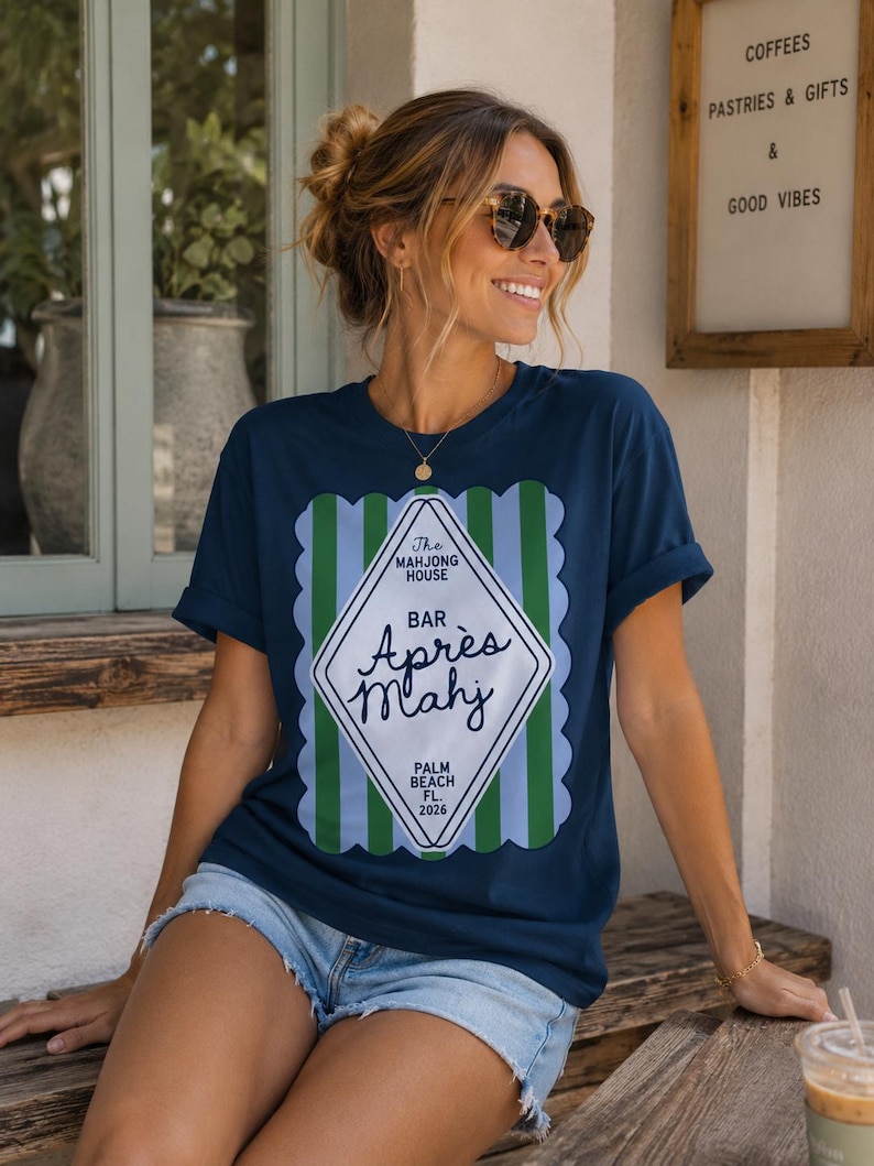 May include: Navy blue t-shirt with a decorative graphic that reads "The Mahjong House Bar Apr&egrave;s Mahj Palm Beach FL. 2026". The shirt has a scalloped border with green and light blue stripes.