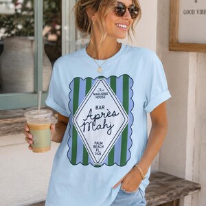 May include: Light blue t-shirt with a graphic that reads "The Mahjong House Bar Apr&egrave;s Mahj Palm Beach FL 2026". The design features a diamond shape with green and blue stripes. The shirt is a casual, everyday wear.