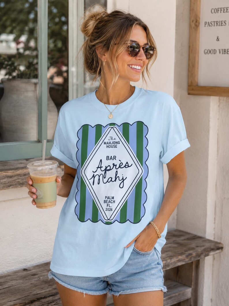 May include: Light blue t-shirt with a graphic that reads "The Mahjong House Bar Apr&egrave;s Mahj Palm Beach FL 2026". The design features a diamond shape with green and blue stripes. The shirt is a casual, everyday wear.