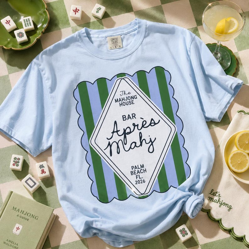 May include: Light blue t-shirt with a diamond-shaped graphic that reads "Apr&egrave;s Mahj" and "The Mahjong House Bar, Palm Beach FL 2026". The shirt is surrounded by mahjong tiles, a book, and a cocktail.