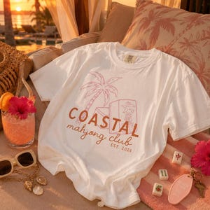 Puede incluir: Camiseta blanca con un gr&aacute;fico rosa y marr&oacute;n que dice "Coastal Mahjong Club EST. 2026." La camiseta est&aacute; sobre una toalla a rayas rosas junto a un c&oacute;ctel, gafas de sol y fichas de mahjong.
