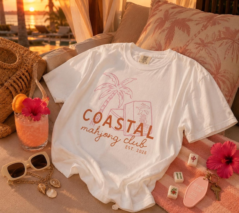 Puede incluir: Camiseta blanca con un gr&aacute;fico rosa y marr&oacute;n que dice "Coastal Mahjong Club EST. 2026." La camiseta est&aacute; sobre una toalla a rayas rosas junto a un c&oacute;ctel, gafas de sol y fichas de mahjong.