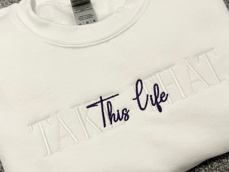 K&ouml;nnte beinhalten: Wei&szlig;es Sweatshirt mit Rundhalsausschnitt und gesticktem Text "TAKE THIS LIFE". Der Text ist in Wei&szlig; und Dunkelblau gehalten und verwendet eine Mischung aus Block- und Schreibschrift. Das Sweatshirt hat eine mittlere Passform.