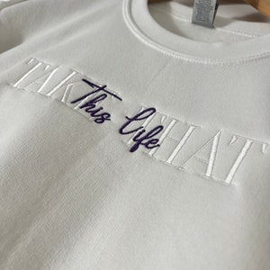 K&ouml;nnte beinhalten: Ein wei&szlig;es Sweatshirt mit dem gestickten Text "TAKE THIS LIFE" in wei&szlig;er und dunkelblauer Farbe. Das Sweatshirt h&auml;ngt an einem Holzb&uuml;gel vor einer wei&szlig;en Wand.