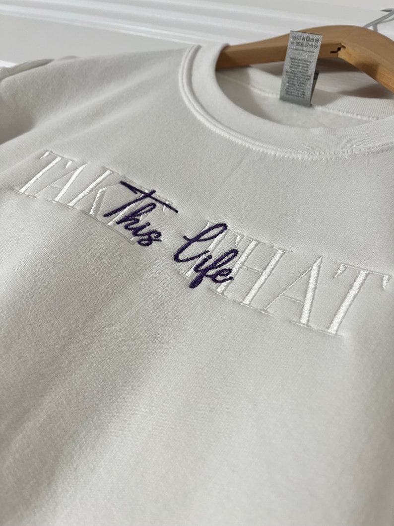 K&ouml;nnte beinhalten: Ein wei&szlig;es Sweatshirt mit dem gestickten Text "TAKE THIS LIFE" in wei&szlig;er und dunkelblauer Farbe. Das Sweatshirt h&auml;ngt an einem Holzb&uuml;gel vor einer wei&szlig;en Wand.