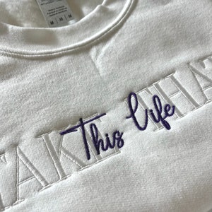 K&ouml;nnte beinhalten: Wei&szlig;es Sweatshirt mit dem gestickten Text "TAKE THIS LIFE" in Lila. Das Sweatshirt hat einen Rundhalsausschnitt und ein Etikett am inneren Kragen. Der Stoff wirkt weich und bequem.